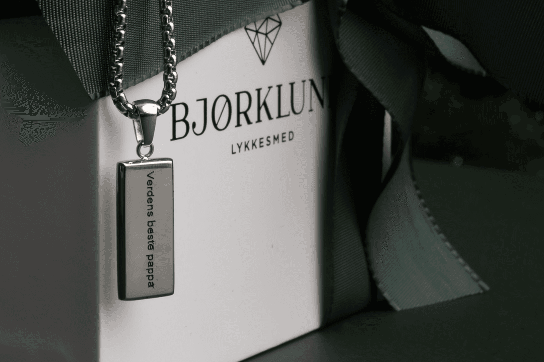 Sølvkjede med et rektangulært anheng gravert med 'Verdens beste pappa' ved siden av en innpakket gaveeske merket 'BJØRKLUND'.