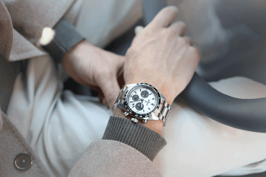 Et tett bilde av fanget til en mann med sort bukse og hvit t-shirt, som folder hendene og viser sin Nixon-klokke.