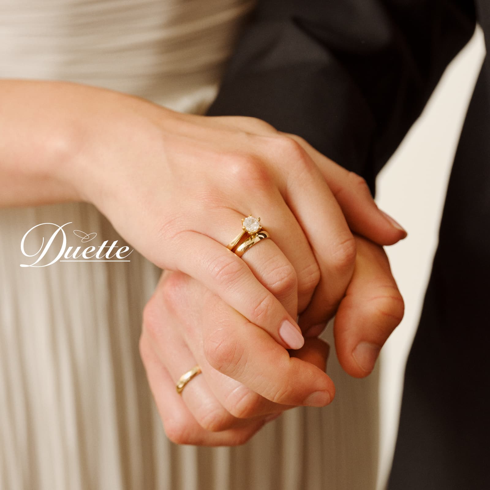 Nærbilde av et par som holder hender, begge med gullringer. Kvinnen har en forlovelsesring med en diamant. Teksten lyder: «Duette.»