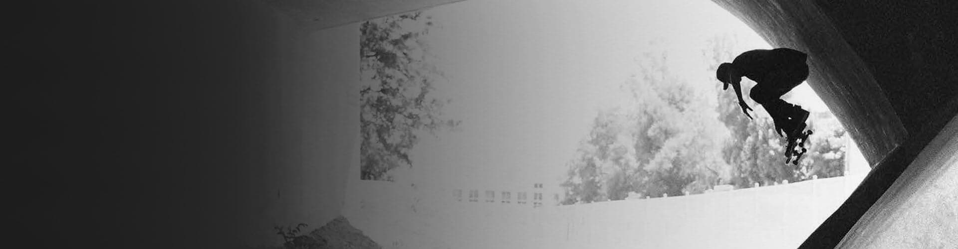 Silhuett av en person som står på skateboard i en betongtunnel og utfører et lufttriks mot et bakteppe av trær og lys himmel.