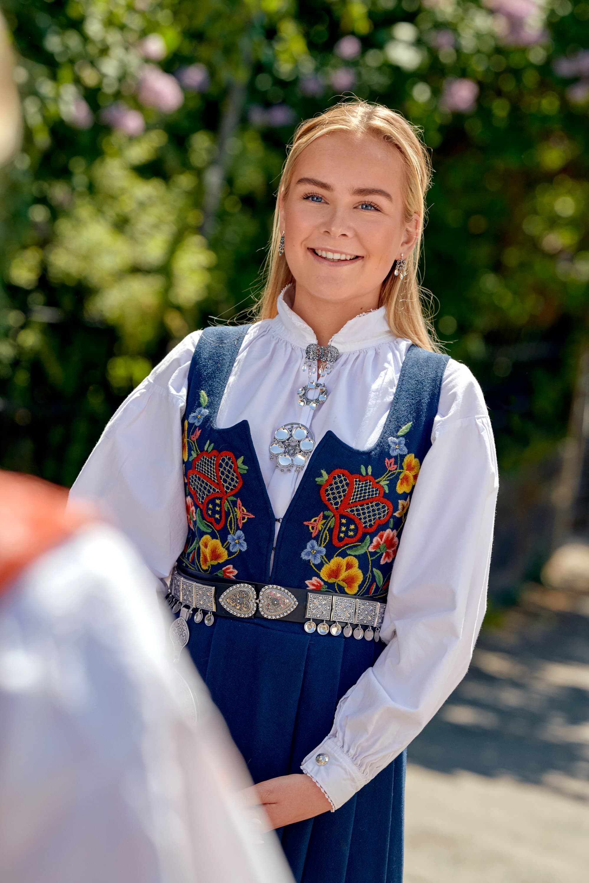 En kvinne i tradisjonell bunad med fargerikt broderi står utendørs og smiler. Hun bærer søljer fra symvsmidja og er omgitt av grønne omgivelser.