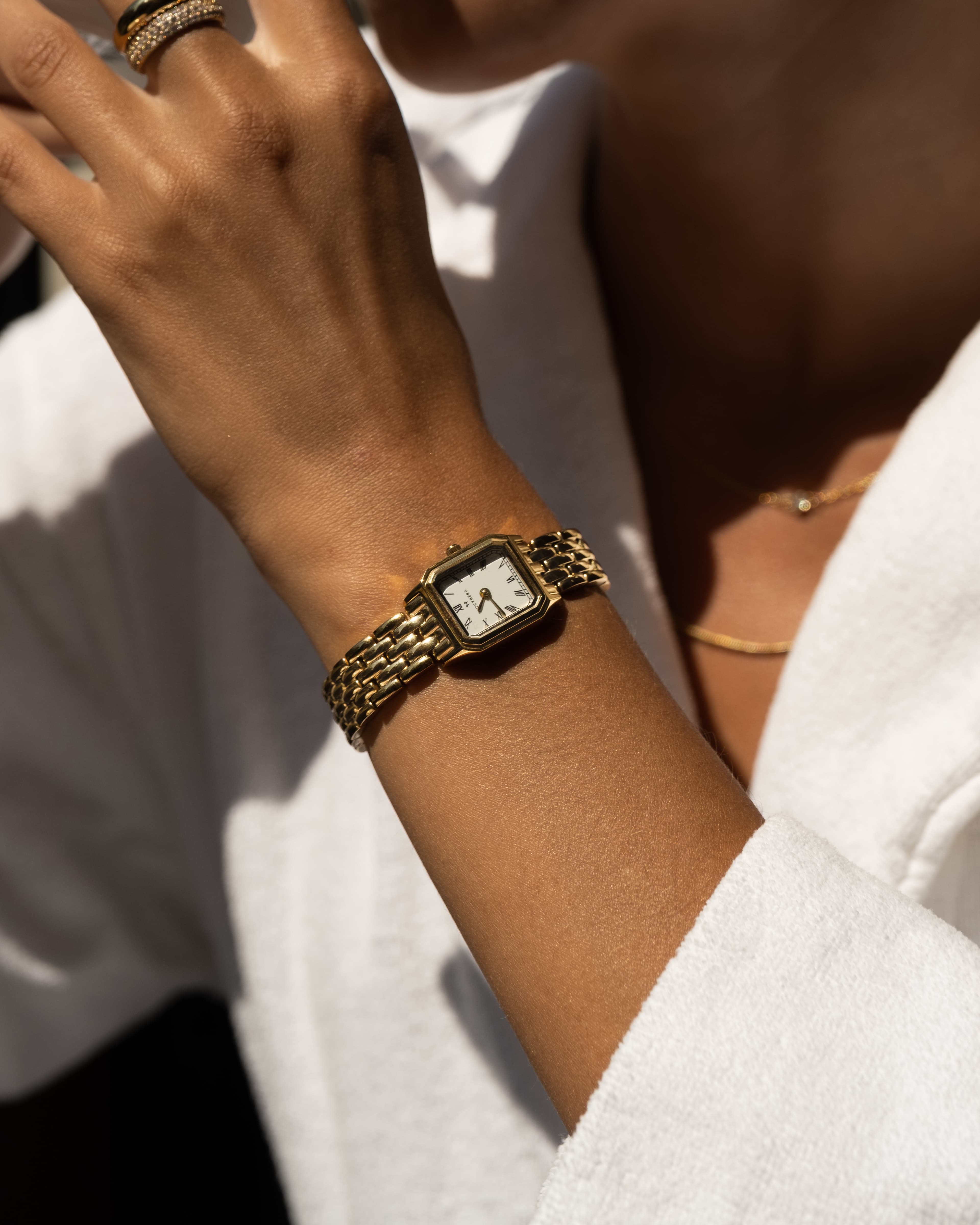 Nærbilde av en person som bærer en gullklokke og en hvit morgenkåpe. Personen har også på seg ringer og et halskjede.