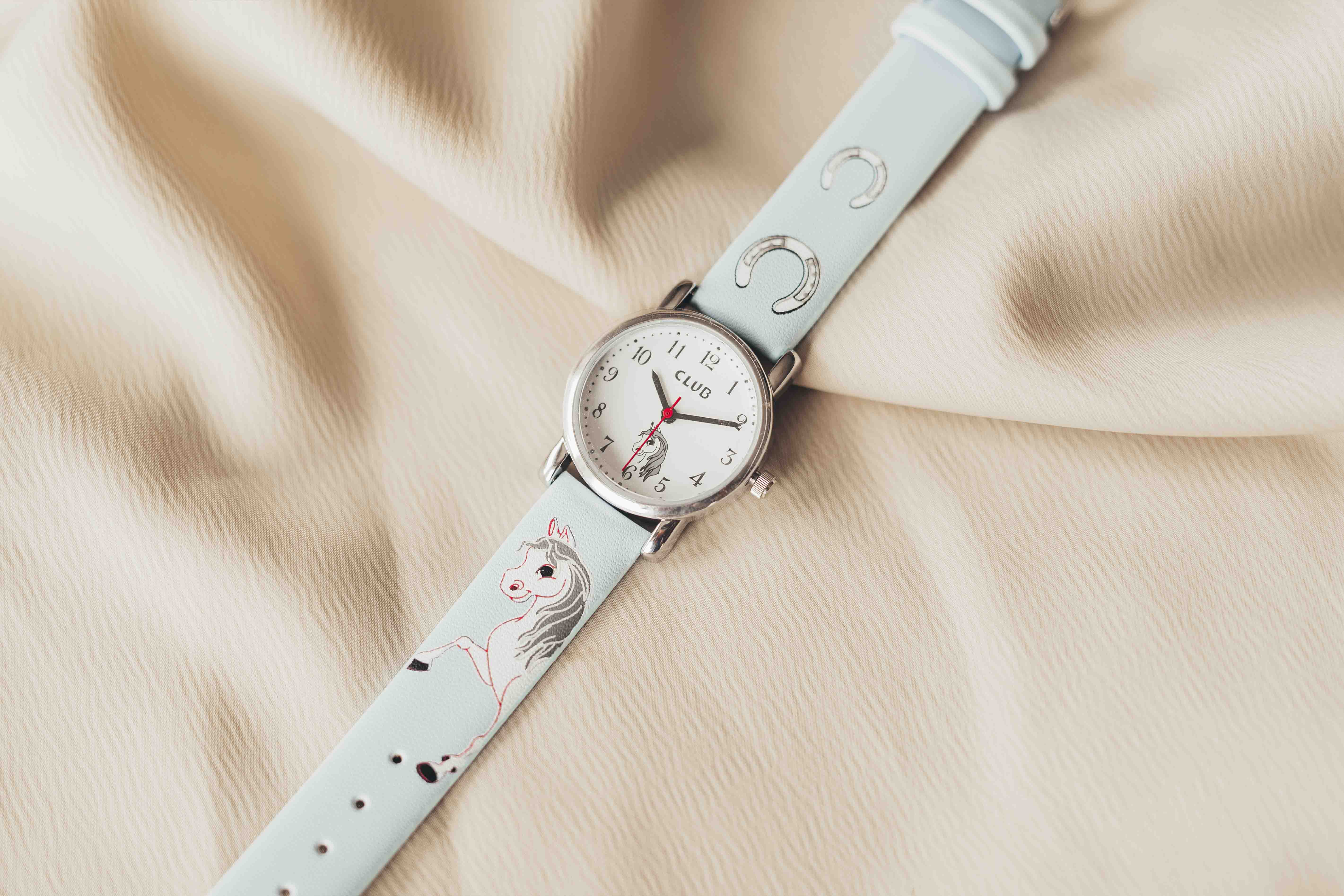 Et lyseblått armbåndsur med hest- og hestesko-design på remmen, hvilende på en myk beige stoffbakgrunn.