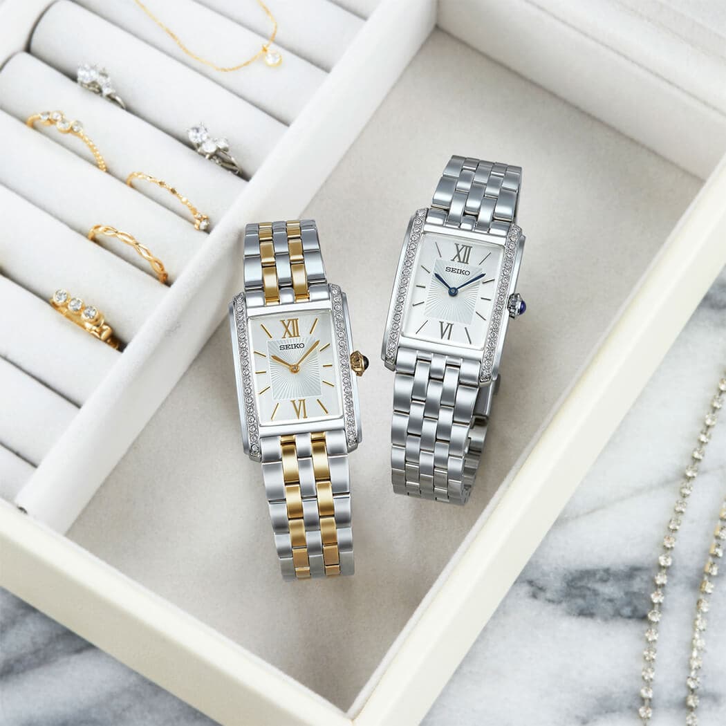 To elegante armbåndsur med rektangulære urkasser, ett i gull og sølv og ett i sølv, ved siden av et smykkeskrin med gull- og sølvgjenstander på en marmoroverflate.