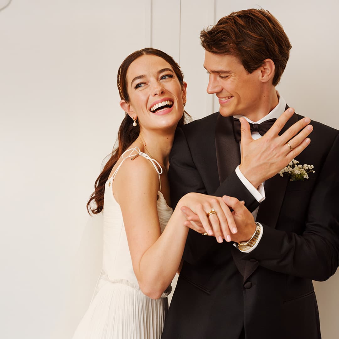 Brud i hvit kjole og brudgom i svart dress deler et gledesfylt øyeblikk, mens de holder hender og smiler mot en hvit bakgrunn.