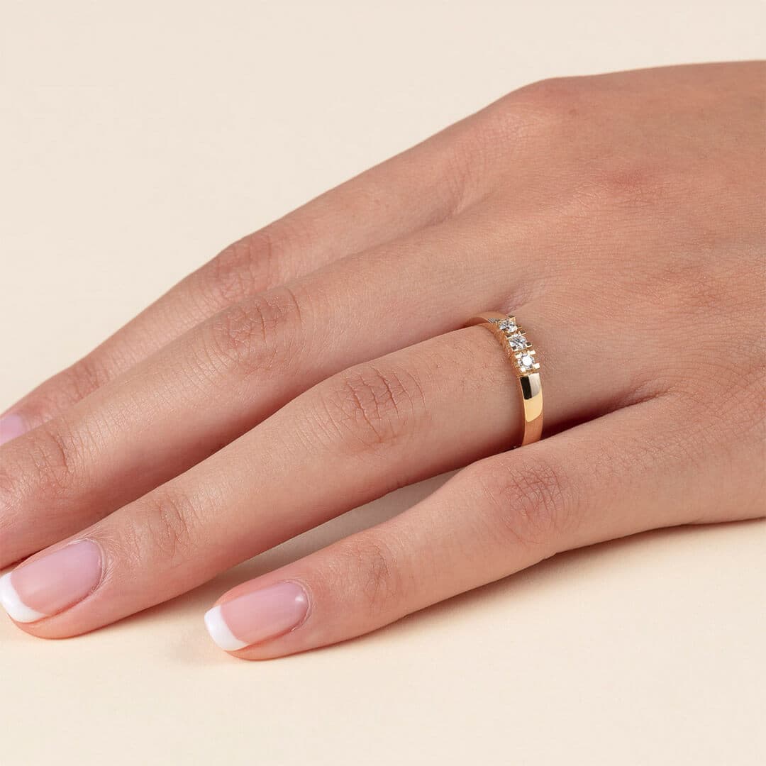 En hånd med fransk manikyr bærer en gullring utsmykket med fem små diamanter på langfingeren, satt mot en myk beige bakgrunn.