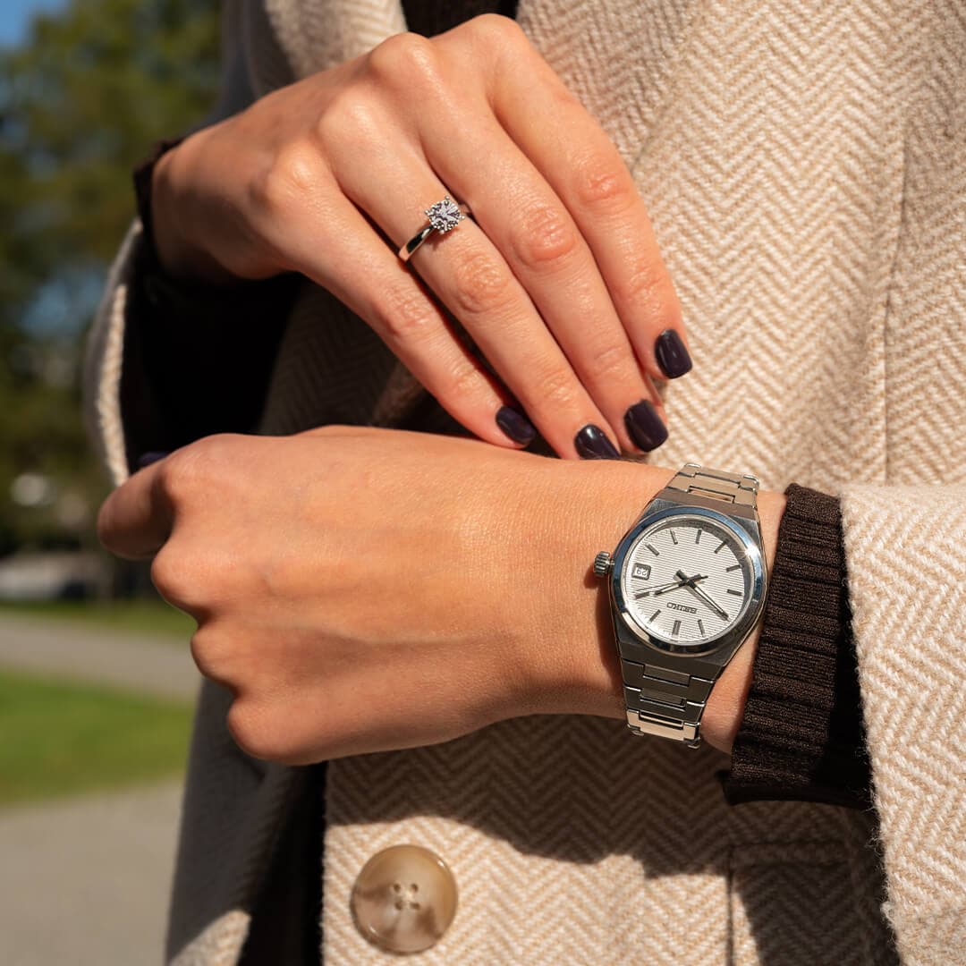 Nærbilde av en person med et sølvfarget armbåndsur og en ring, med mørk neglelakk, i en beige frakk utendørs.