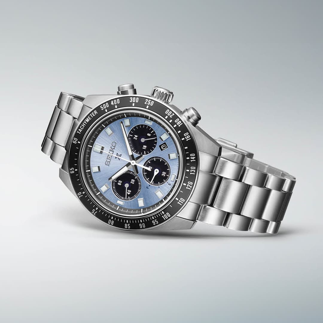 Sølvfarget kronografklokke med blå urskive, to svarte underurskiver, takymeterskala på bezel og metalllenke.