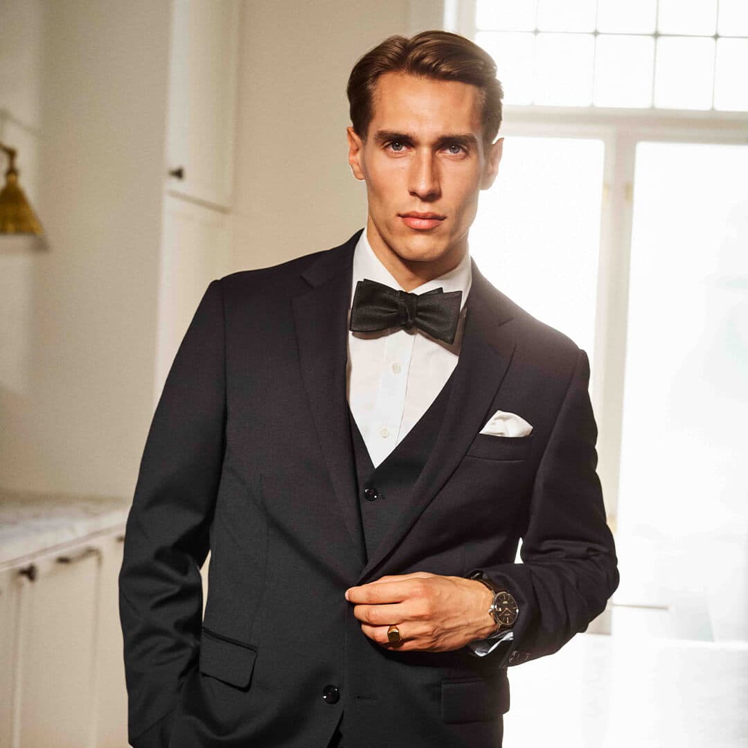 Mann i svart dress med sløyfe, stående selvsikkert i et solfylt rom med hvite vegger og et vindu i bakgrunnen.