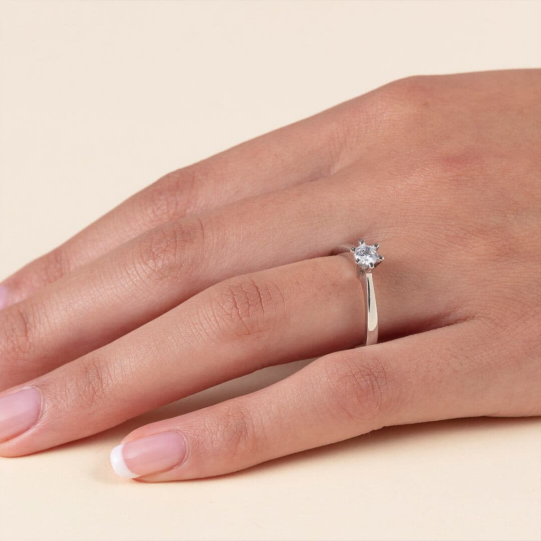 Nærbilde av en hånd med en forlovelsesring i sølv med en rund solitærdiamant, mot en beige bakgrunn.