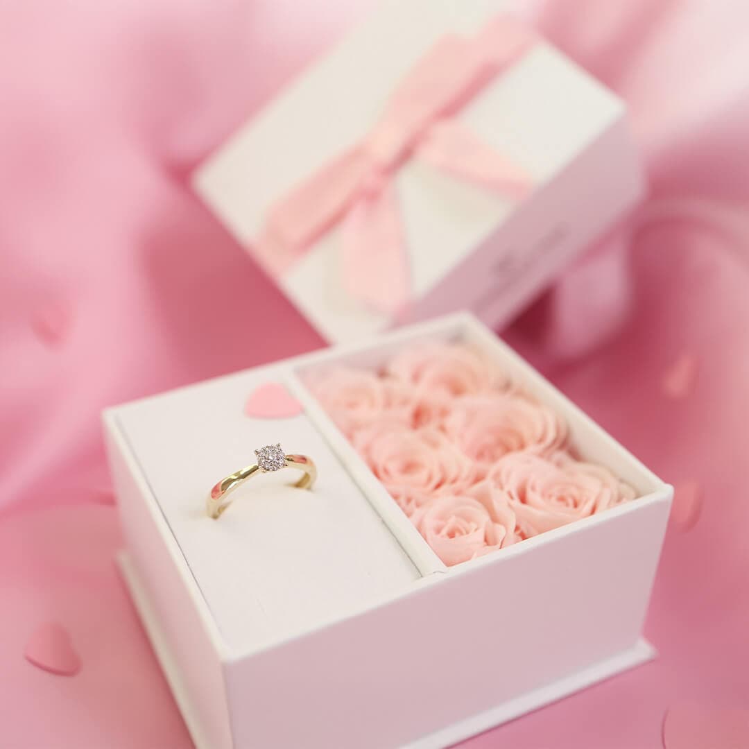 Åpen eske med en gullforlovelsesring og en del av rosa roser på en rosa stoffbakgrunn.