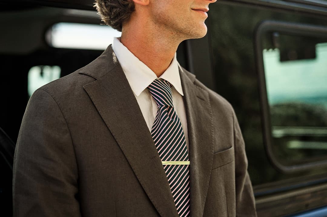 Mann i brun dress og stripete slips stående ved siden av et kjøretøy, delvis synlig.