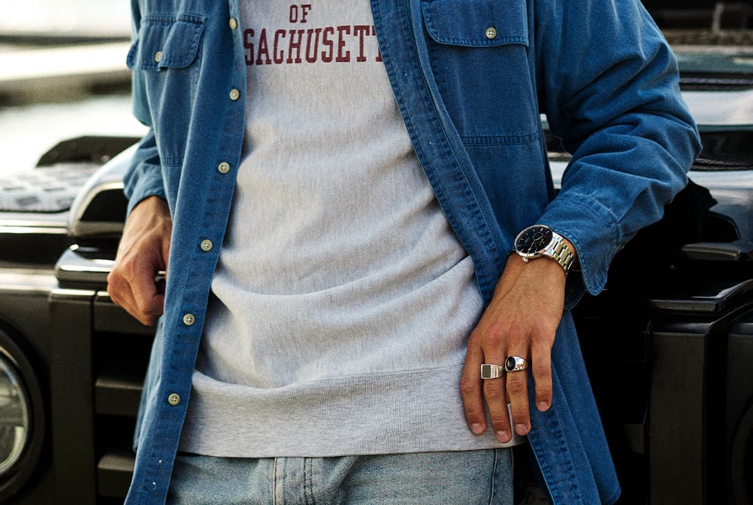 Person iført en blå denimskjorte, grå genser med teksten 'Massachusetts', jeans og ringer, stående ved et svart kjøretøy.