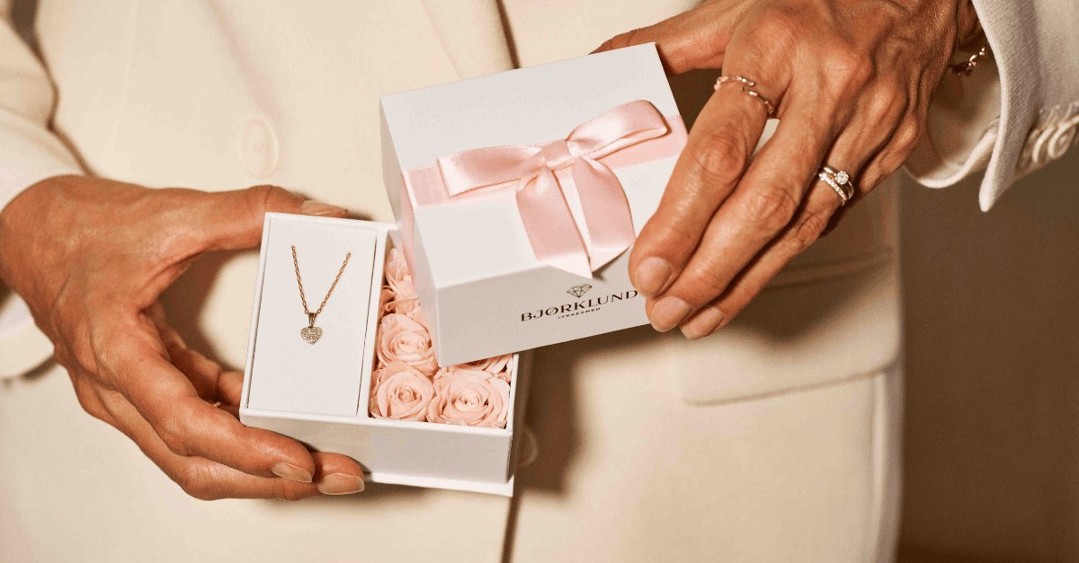 En kvinne med rosa kjole holder en hvit gaveeske med rosa bånd på utsiden og rosa roser på innsiden