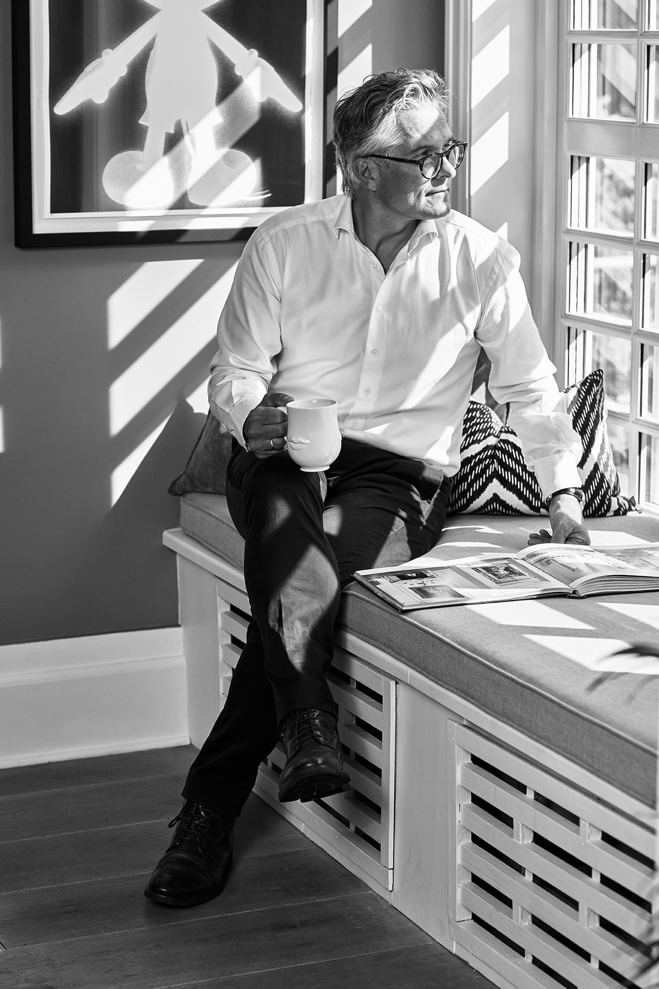 Mann i hvit skjorte og briller sitter på en polstret benk, holder en kopp og ser ut av et solbelyst vindu ved siden av et magasin og et kunstverk.