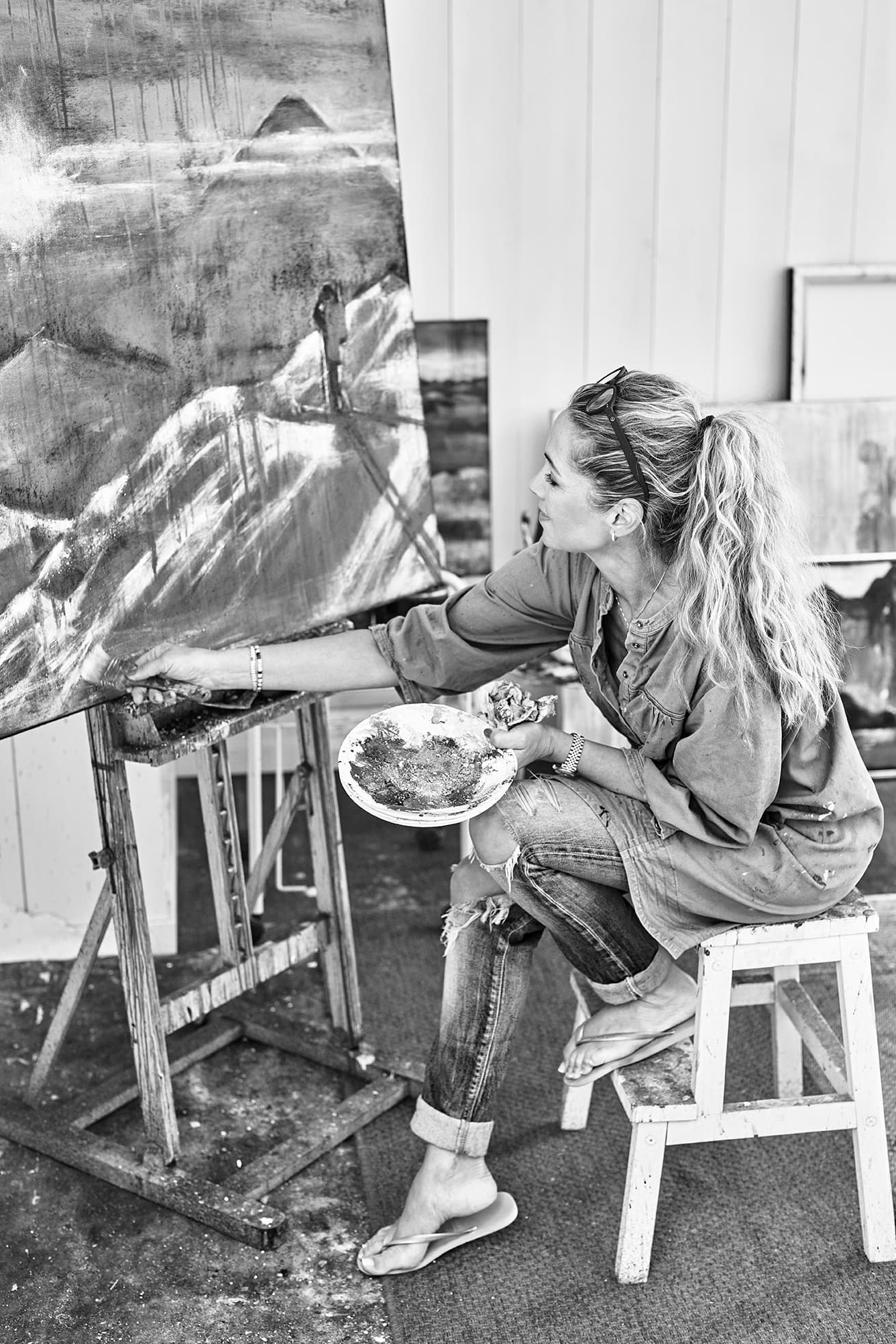 Kunstner i et atelier som maler på et stort lerret, sittende på en krakk. Hun holder en palett og har på seg uformelle klær, omgitt av andre kunstverk.