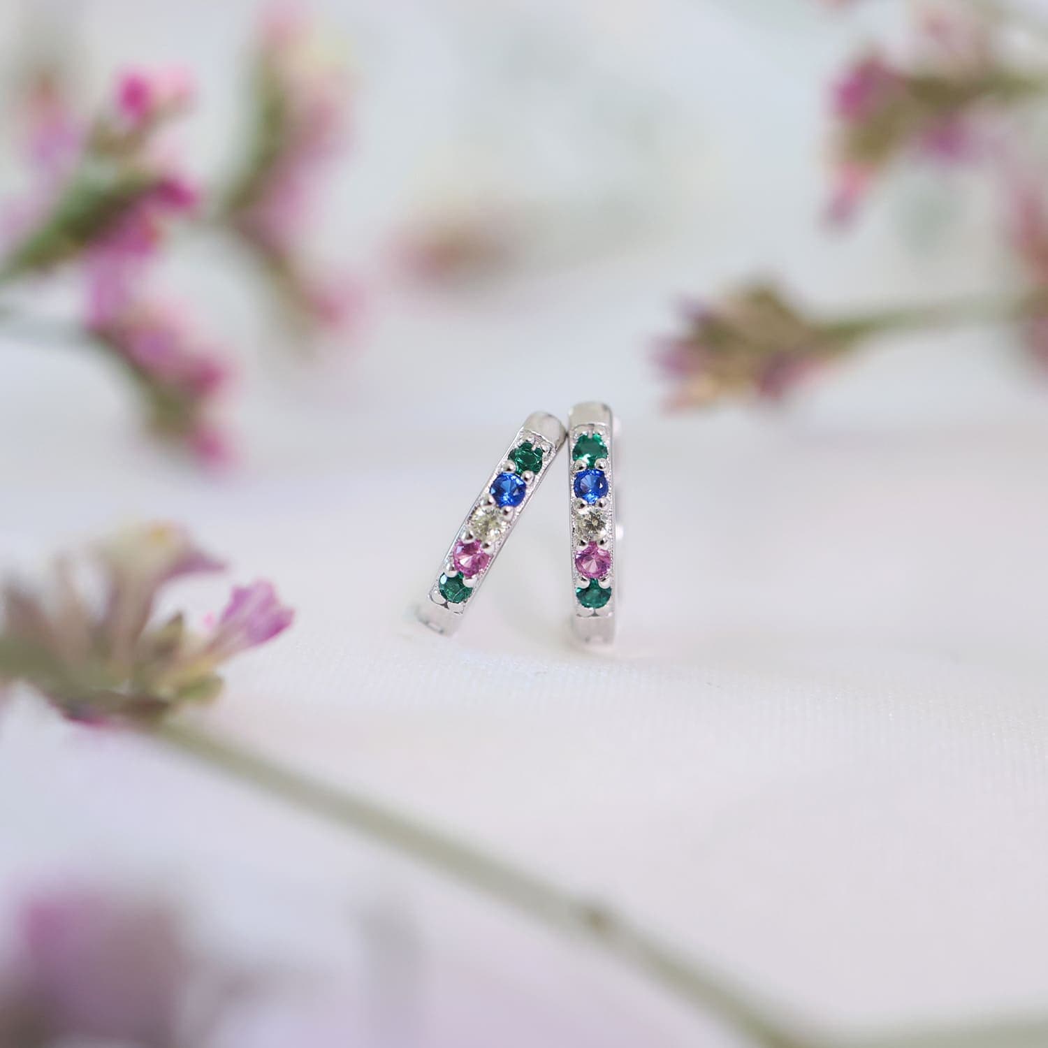 Sølvfargede kreoler med flerfargede edelstener plassert på en myk hvit overflate, omgitt av uklare rosa blomster.