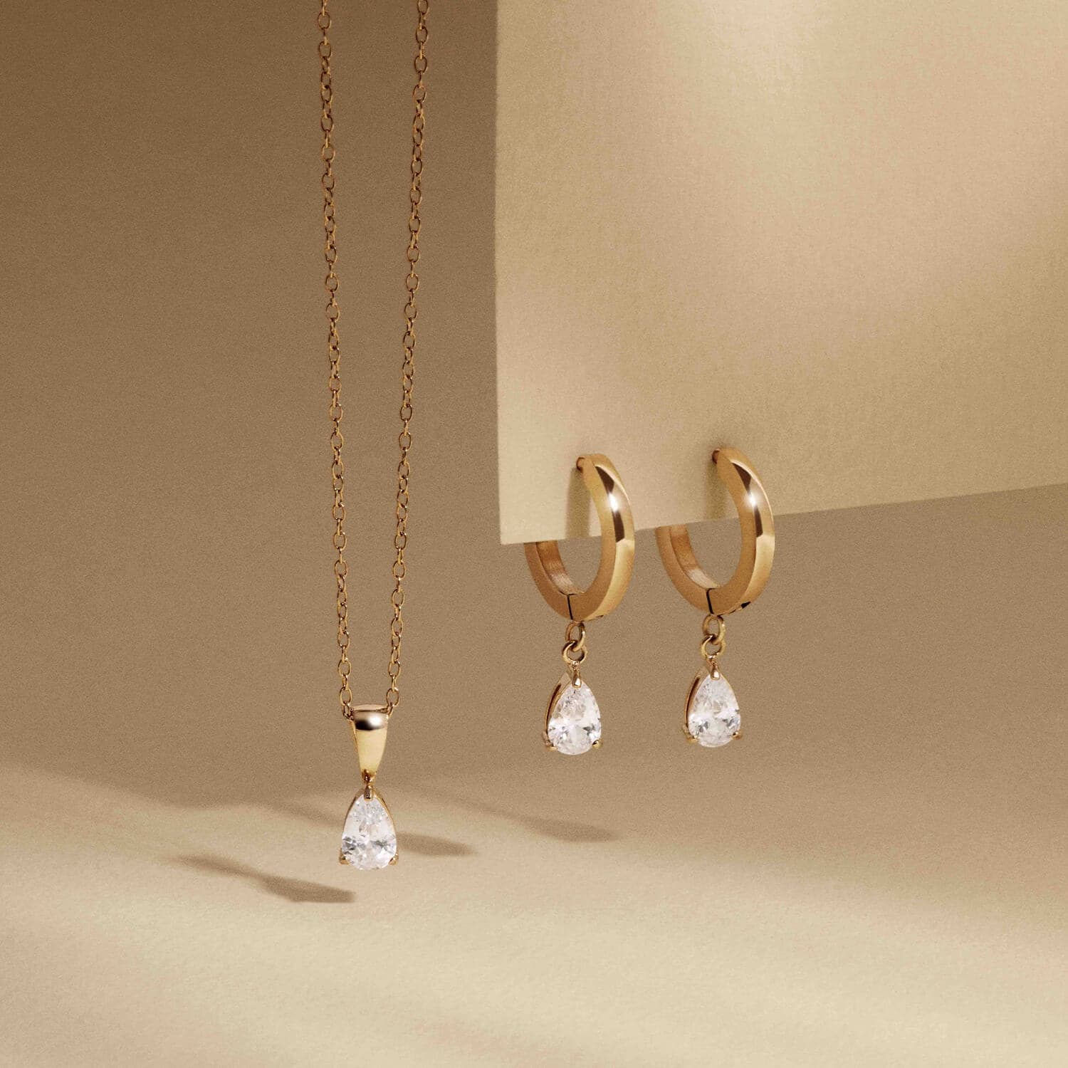 Gullkjede med dråpeformet anheng og matchende gullfargede øredobber, hver med dinglende dråpeformede krystaller, mot en beige bakgrunn.