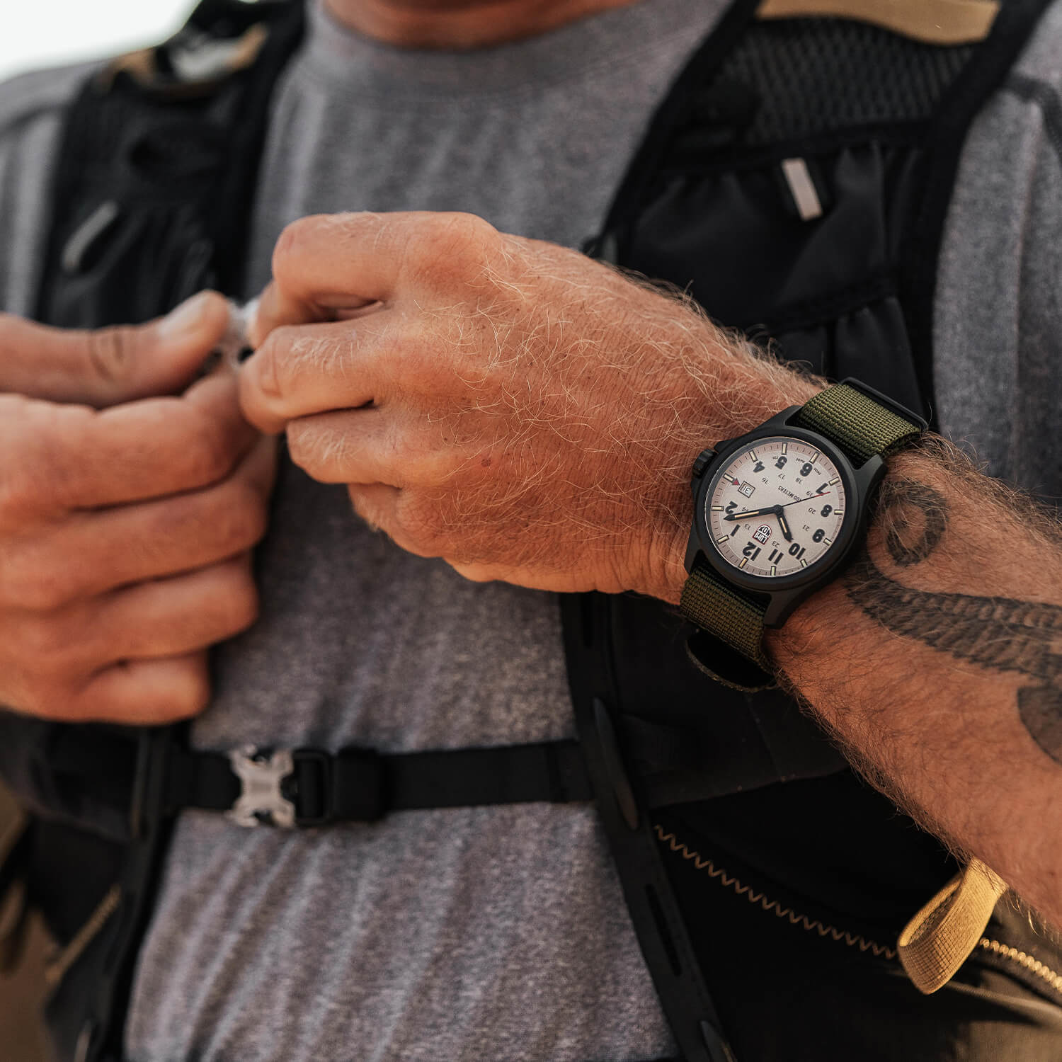 Person som retter på en svart taktisk vest, iført en grønn armbåndsur med hvit urskive og en synlig tatovering på underarmen.