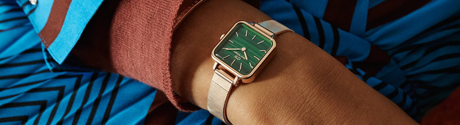 Et håndledd prydet med en roségullklokke med en grønn rektangulær urskive og nettingrem, mot en bakgrunn av blått og brunt mønstret stoff.