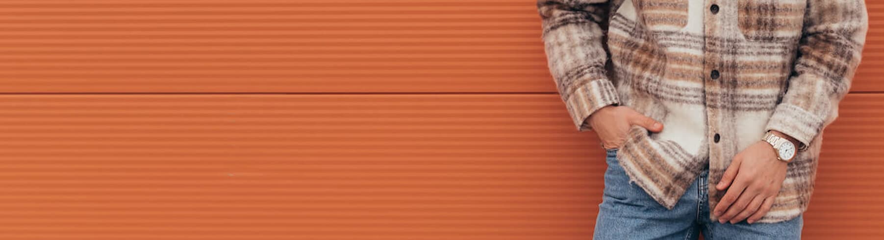 En person i rutete jakke og jeans står mot en oransje rillet vegg med hendene i lommene og en klokke synlig på håndleddet.
