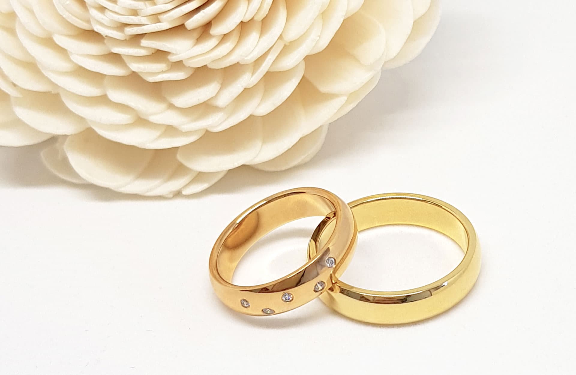 To gullringer, den ene med små diamanter, plassert ved siden av en stor, hvit blomst med tekstur på en lys overflate.