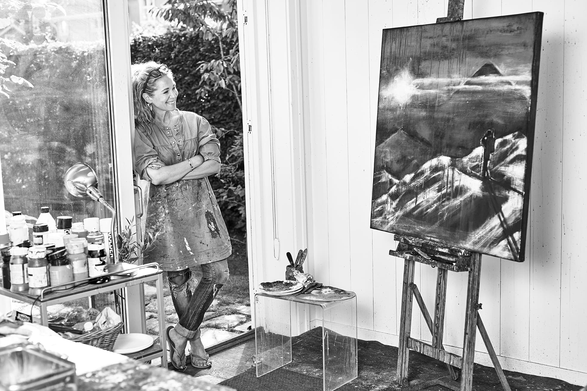 Kunstner i et atelier som betrakter et landskapsmaleri på et staffeli, omgitt av kunstmateriell, med et stort vindu som viser grøntområder utenfor. Svart-hvitt.
