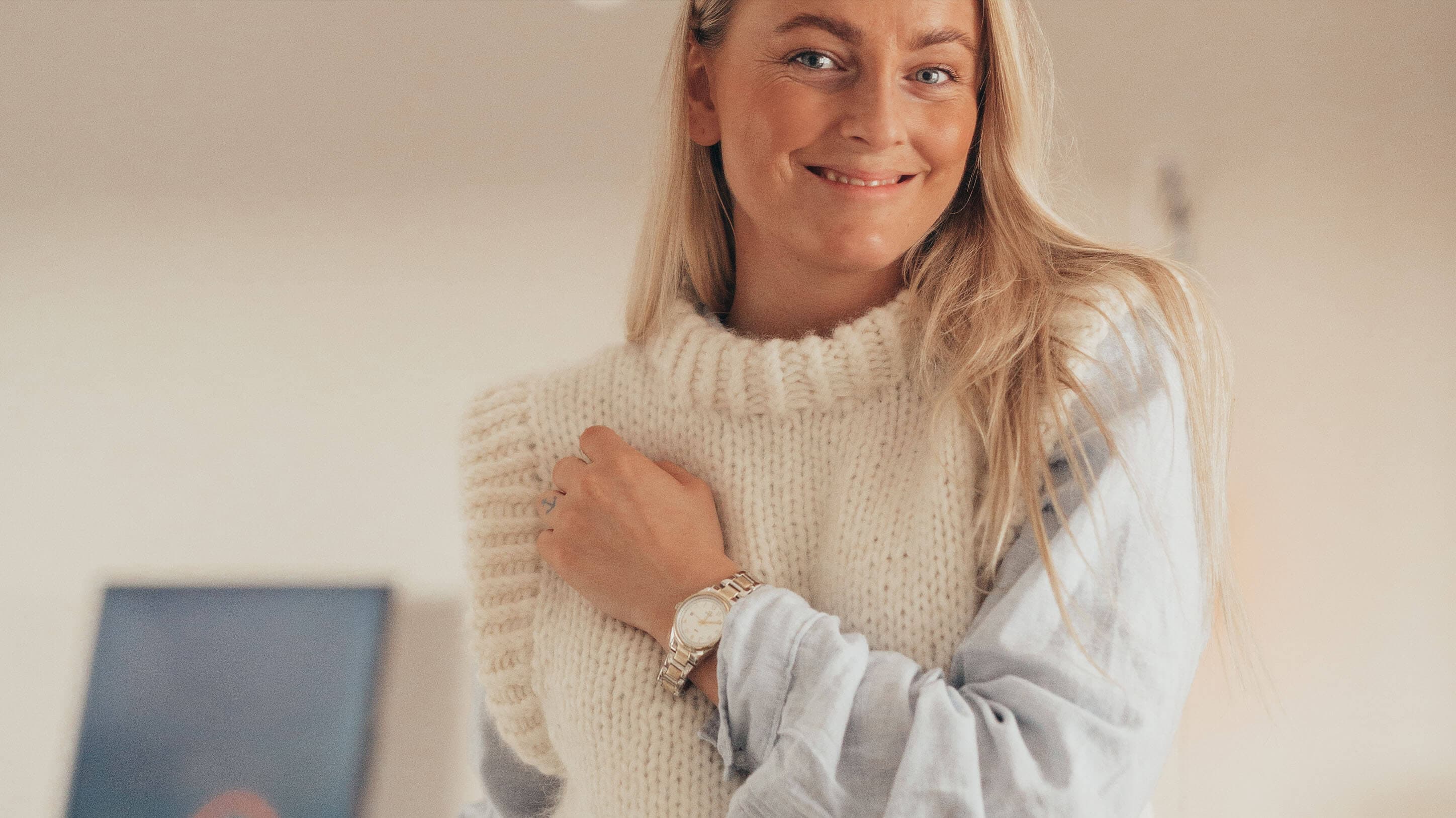 Smilende kvinne med langt blondt hår iført en tykk strikket genser over en lyseblå skjorte, med et armbåndsur. Nøytral bakgrunn.