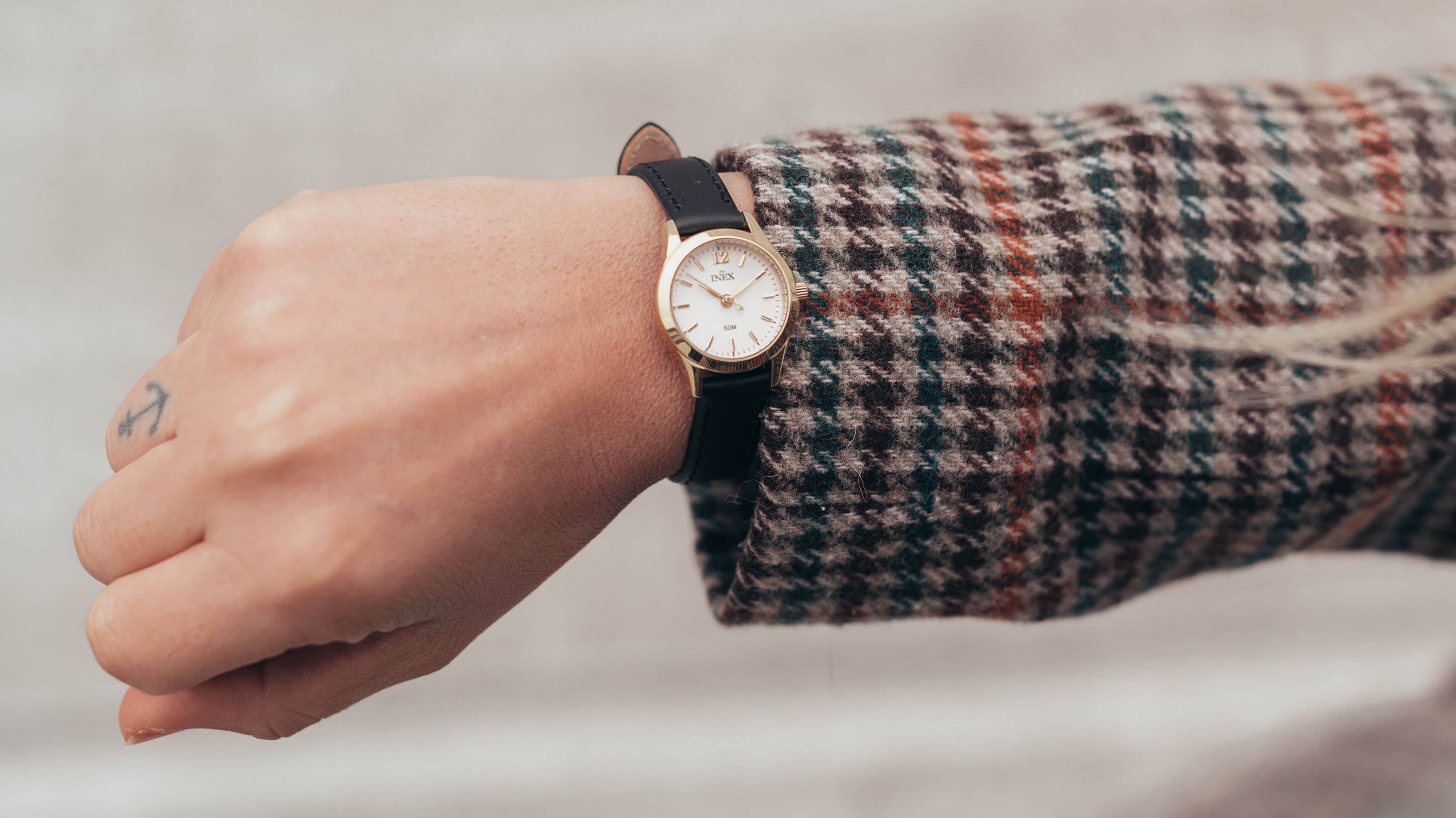 En hånd iført en rutete jakke og et armbåndsur med svart lærreim og gullurkasse.