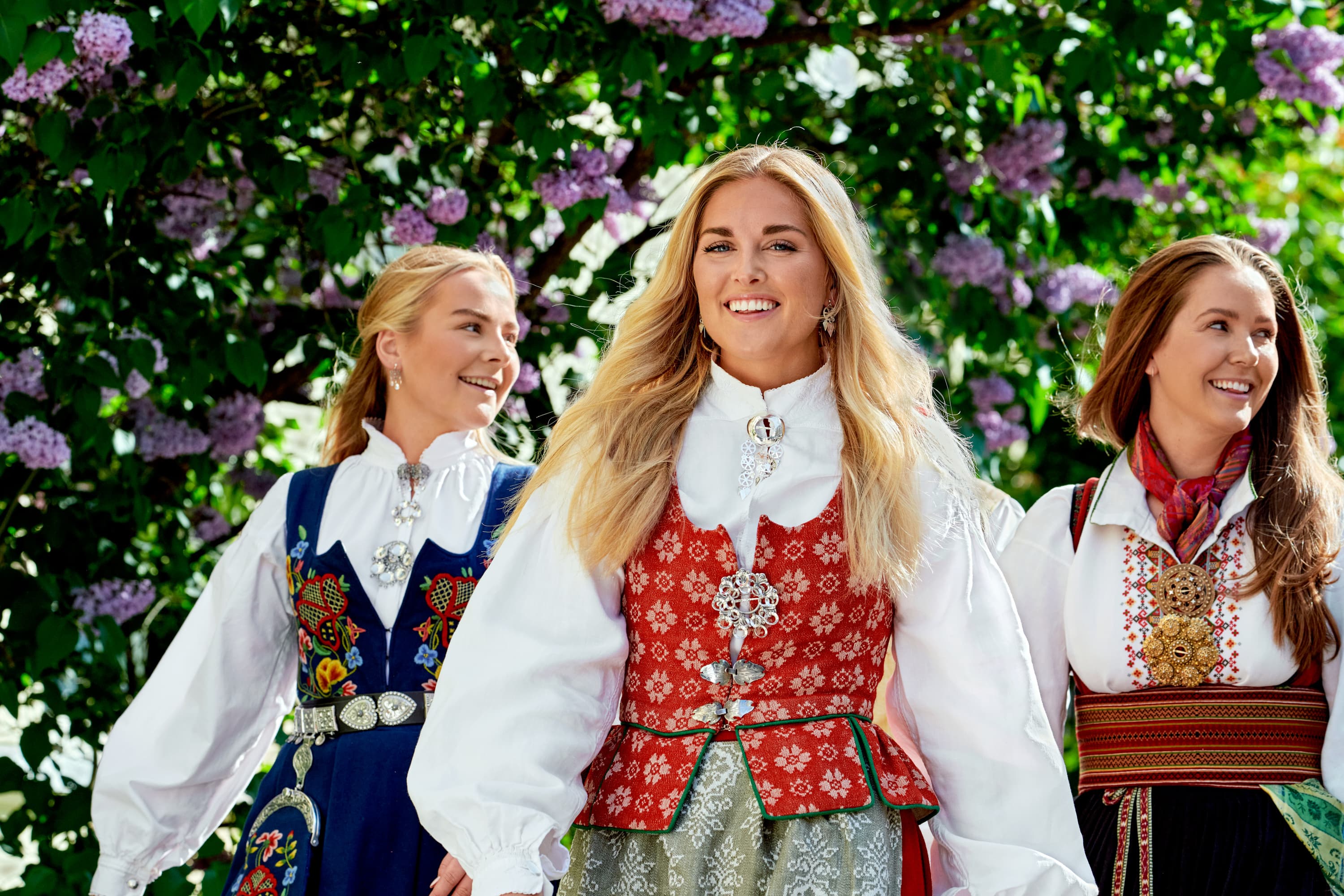 Tre kvinner i fargerike bunader går gladelig foran blomstrende syrinbusker, smilende og nytende en solrik dag.