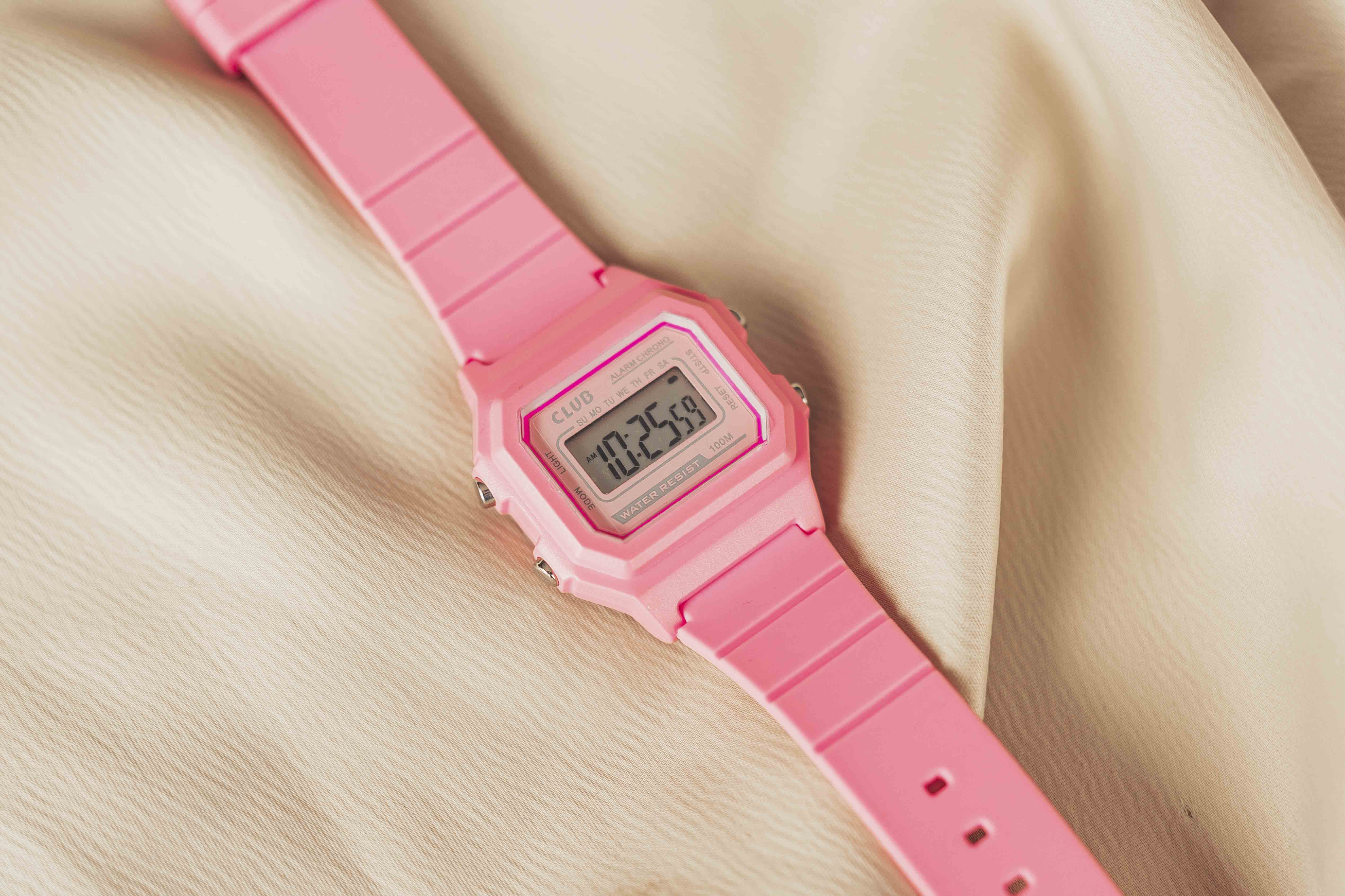 Rosa digital armbåndsur med firkantet urskive og rosa rem på beige stoffbakgrunn. Skjermen viser tiden med knapper på sidene.