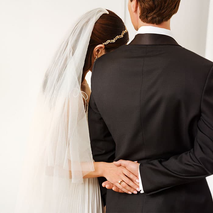 Bruden lener seg på brudgommens skulder, armen hennes knyttet gjennom hans, begge vendt bort. Bruden bærer slør; brudgommen i dress.