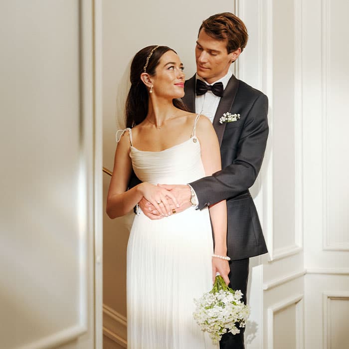 En brudgom i dress omfavner sin brud i hvit kjole som holder blomster, mens de står i et rom med myk belysning og ser kjærlig på hverandre.