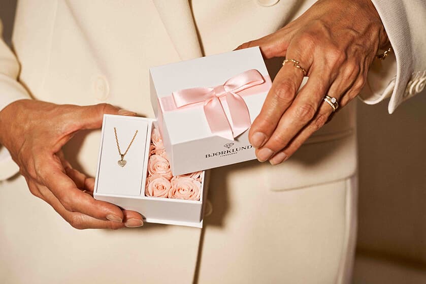 En kvinne i hvit dress holder en Bjørklund-gaveeske med et hjerteanheng og små rosa roser. Esken har en rosa silkesløyfe.