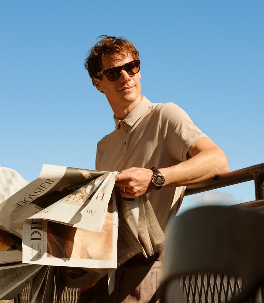 En mann i solbriller står utendørs, holder en avis og lener seg mot et rekkverk, under en klar blå himmel.