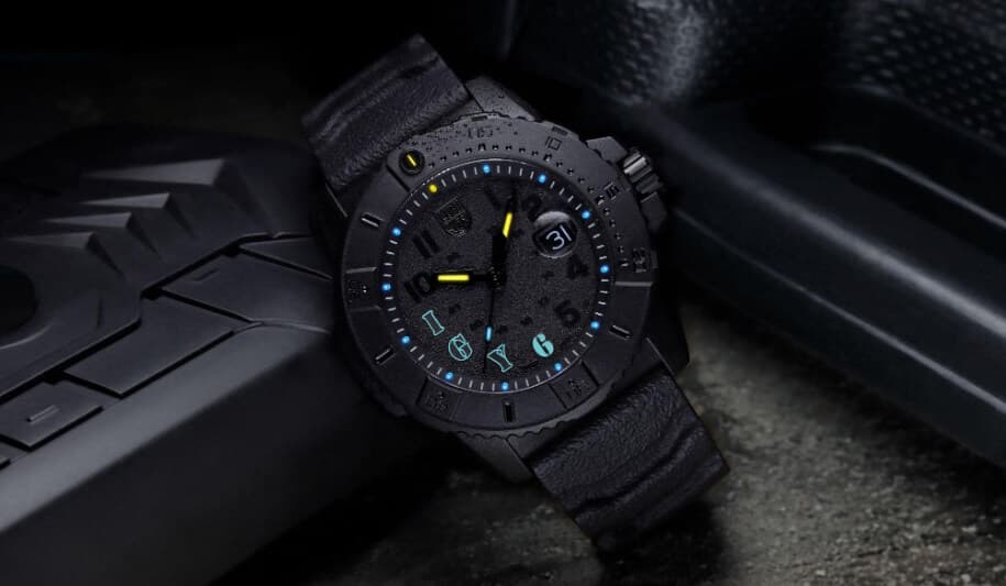 Et robust svart armbåndsur med teksturert bånd, blå timemarkører og datovisning, hvilende på en mørk overflate.