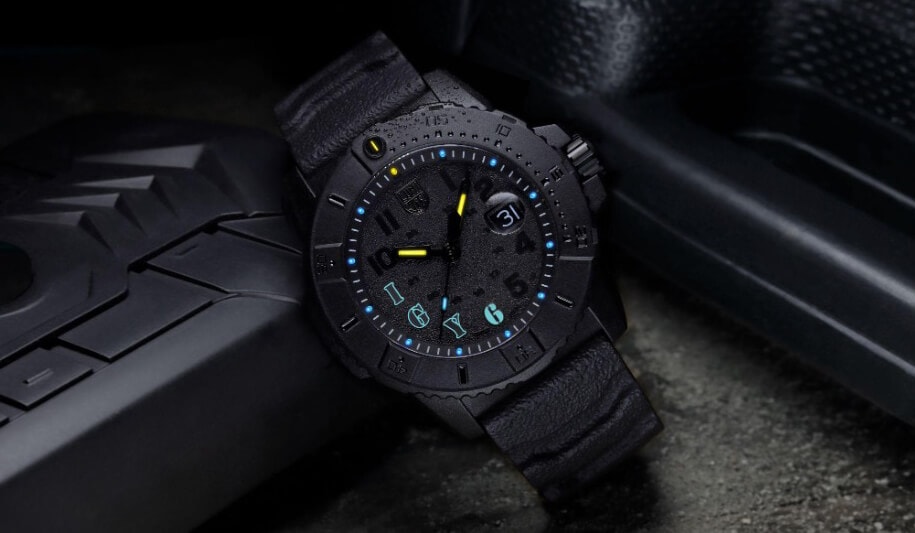Et robust svart armbåndsur med teksturert bånd, blå timemarkører og datovisning, hvilende på en mørk overflate.