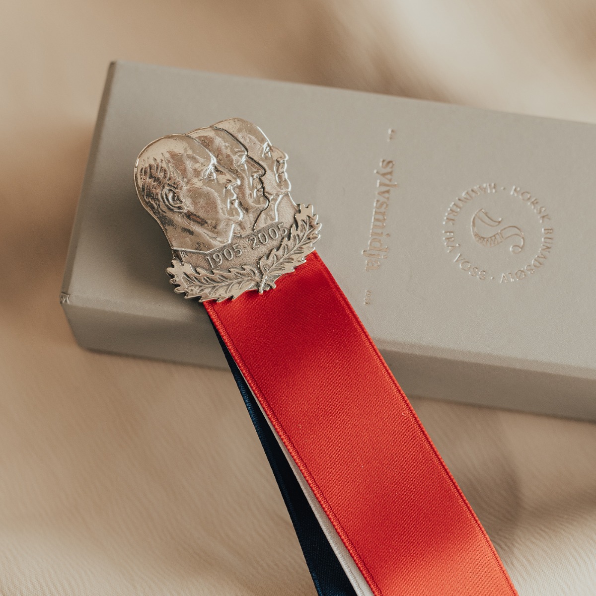 Sølvsløyfe med motiv av tre profiler og hengende bånd i rødt, hvitt og blått som symboliserer Norge.