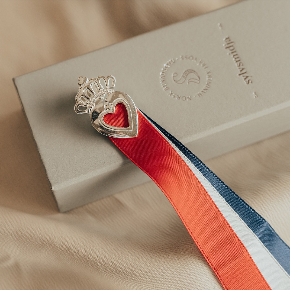 Sølvsløyfe med rødt hjerte og krone øverst, og rødt, hvitt og blått bånd hengende ned mot hvit bakgrunn