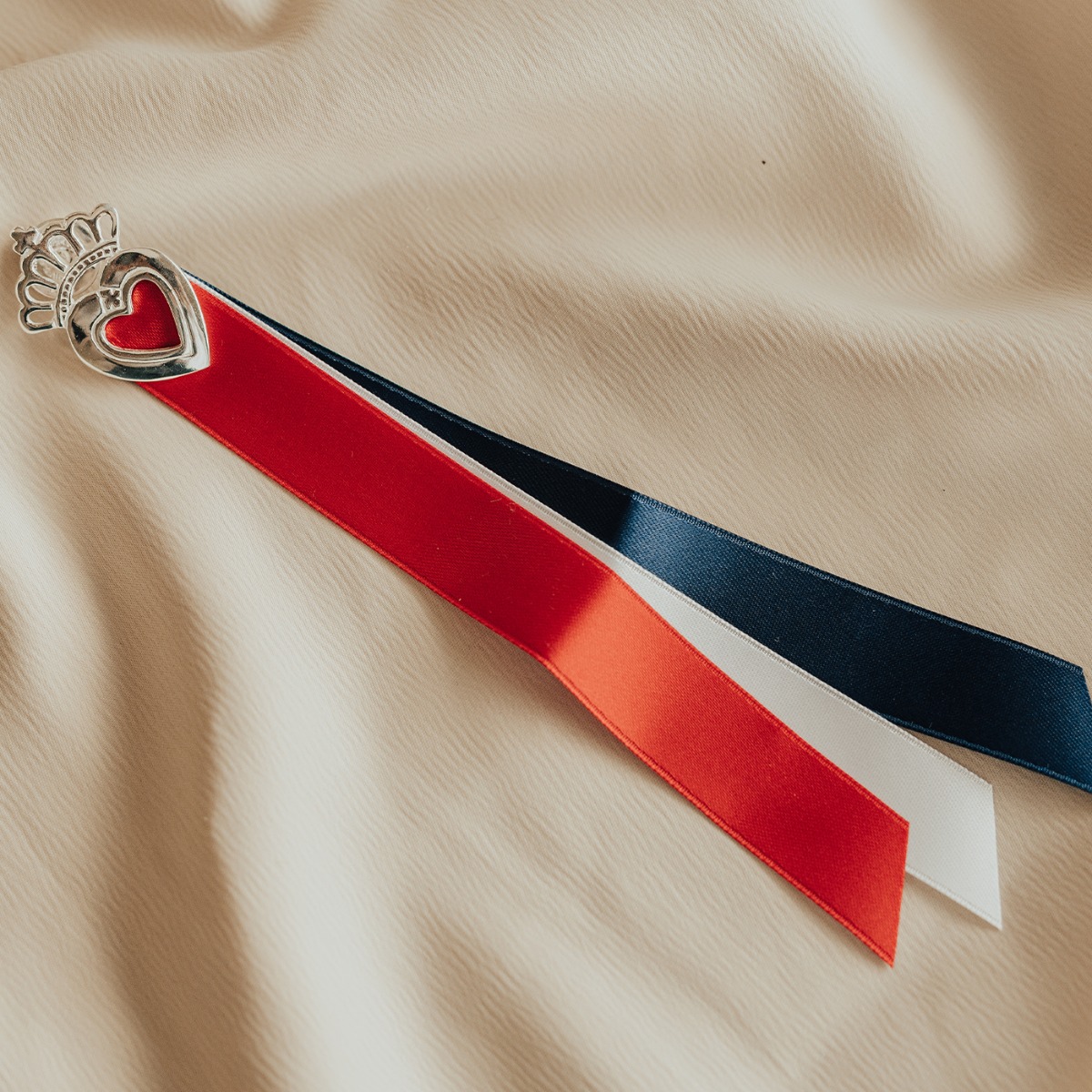 Sølvsløyfe med rødt hjerte og krone øverst, og rødt, hvitt og blått bånd hengende ned mot hvit bakgrunn