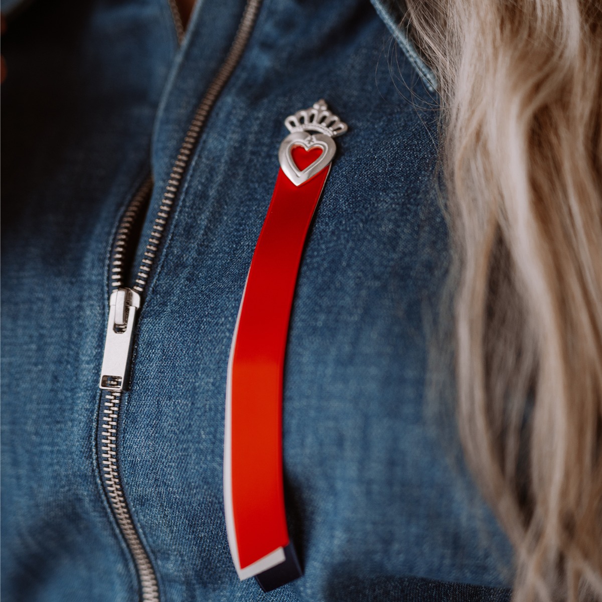 Sølvsløyfe med rødt hjerte og krone øverst, og rødt, hvitt og blått bånd hengende ned mot hvit bakgrunn