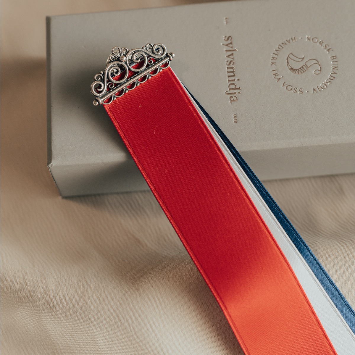 Rød, hvit og blå sløyfe med sølvfarget filigrandekor øverst, avbildet mot hvit bakgrunn.