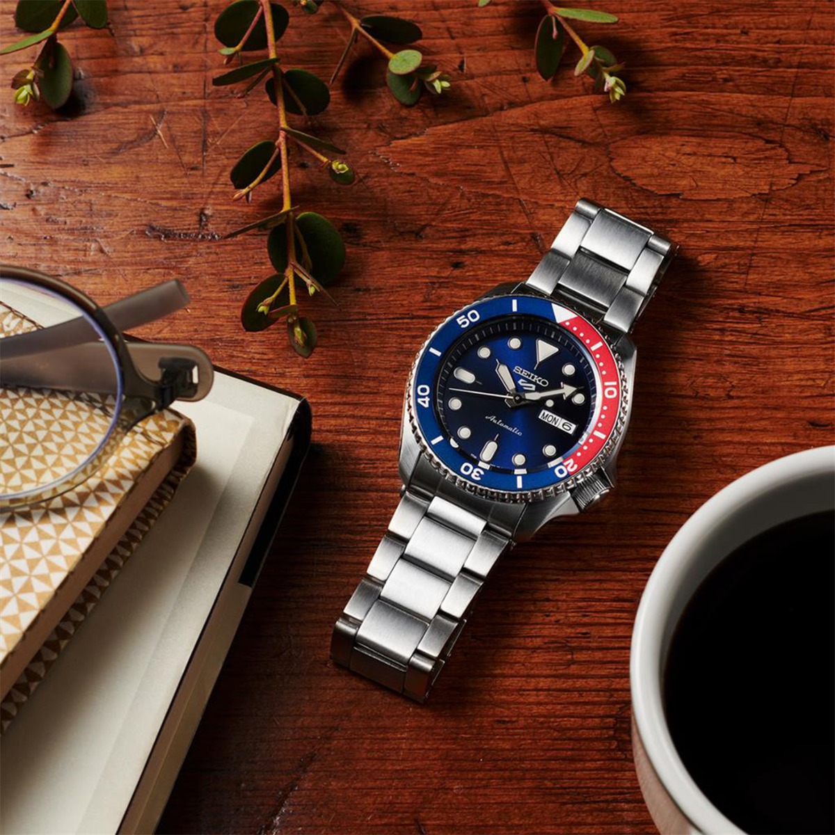 Dykkeklokke i rustfritt stål med mørkeblå urskive, tofarget rød og blå roterende bezel, dag-dato-vindu og stållenke mot hvit bakgrunn