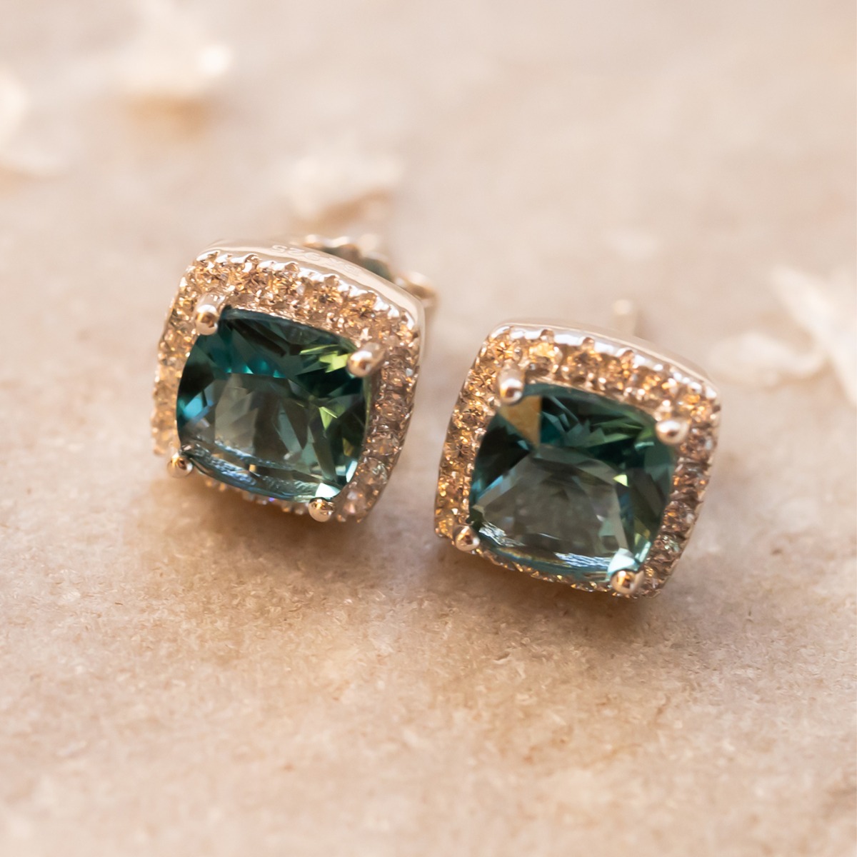 To sølvfargede øredobber med firkantede blå steiner omgitt av små klare stener i en blank innfatning.