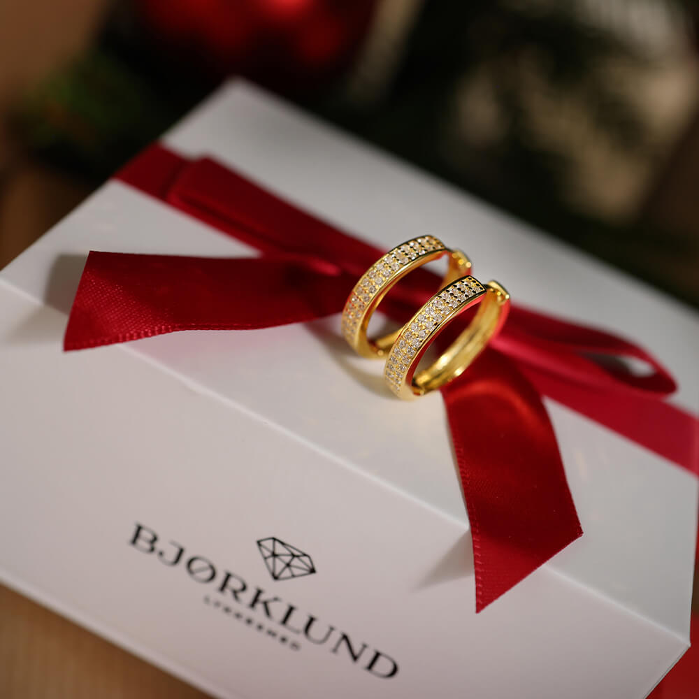 Forgylte øreringer i sølv med brede bånd, tett besatt av små klare stener på fremsiden, vist mot hvit bakgrunn.