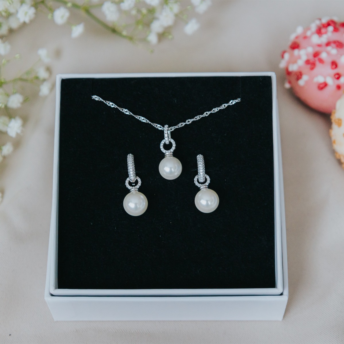 Par med sølvfargede hengende øredobber med store hvite perler og små klare steiner på bøylene, avbildet mot hvit bakgrunn