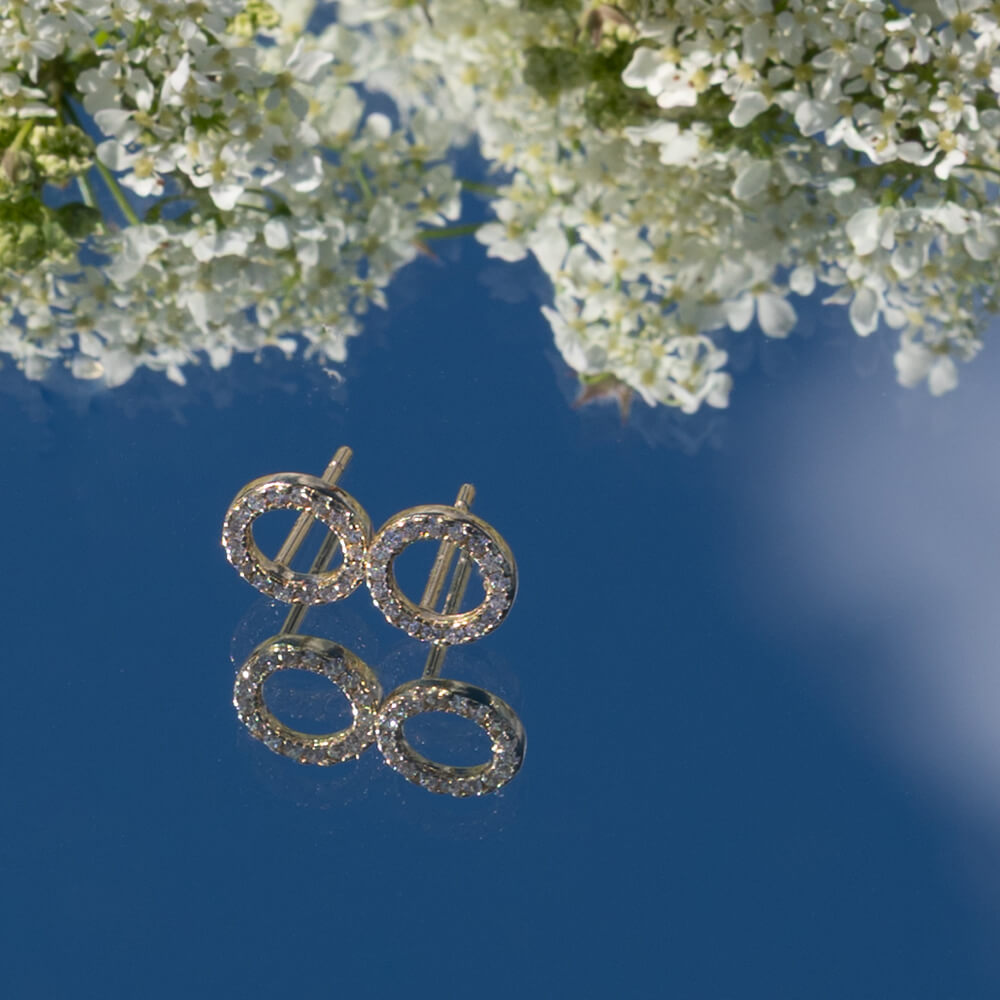 To små, sirkelformede øredobber i gull med klare steiner, plassert mot hvit bakgrunn med svak refleksjon under.