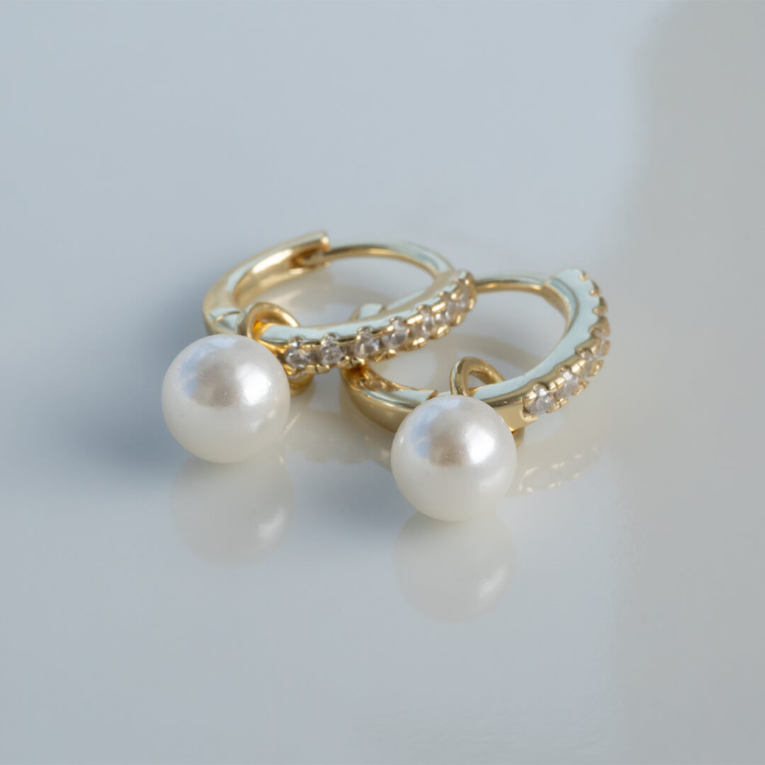 Par gullbelagte creol-øredobber med gjennomsiktige steiner på utsiden og hvite perler hengende fra innsiden mot hvit bakgrunn