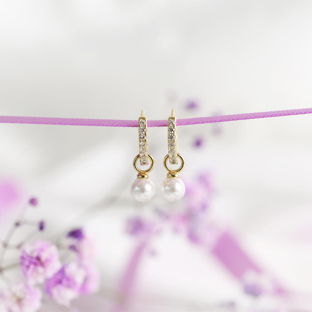 Par gullbelagte creol-øredobber med gjennomsiktige steiner på utsiden og hvite perler hengende fra innsiden mot hvit bakgrunn