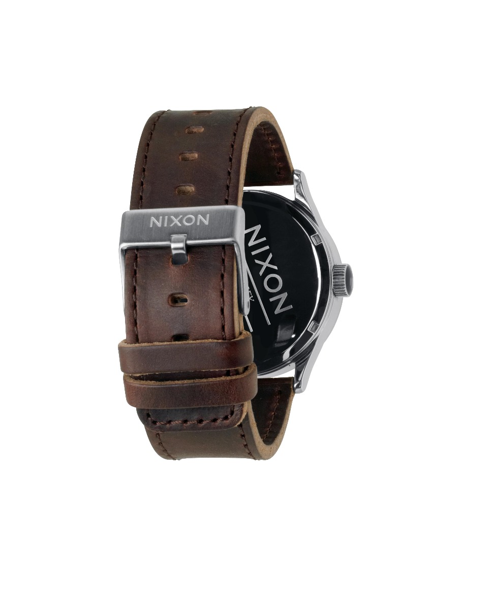 Sølvfarget Nixon-klokke med hvit skive, røde stavindekser og sølv visere, dag-dato vindu og mørkebrun lærrem med søm